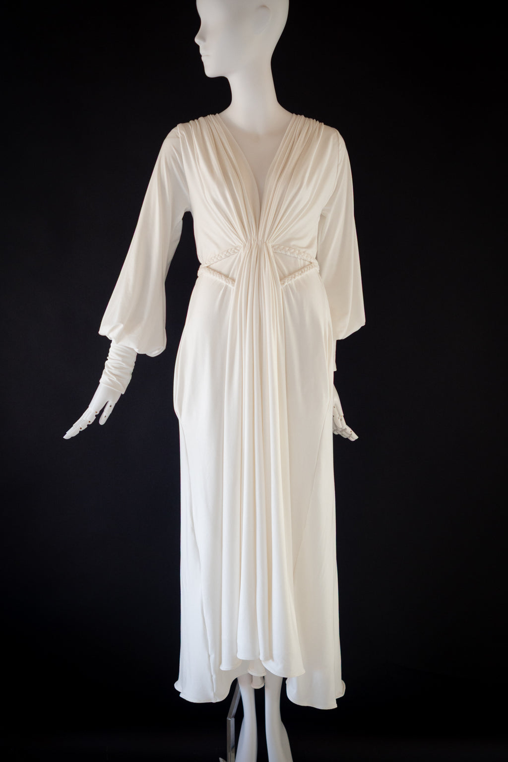 An off-white goddess style draped jersey gown displayed on a white mannequin against a solid black background. The dress features a gathered waist, three-quarter sleeves, and a draped neckline.