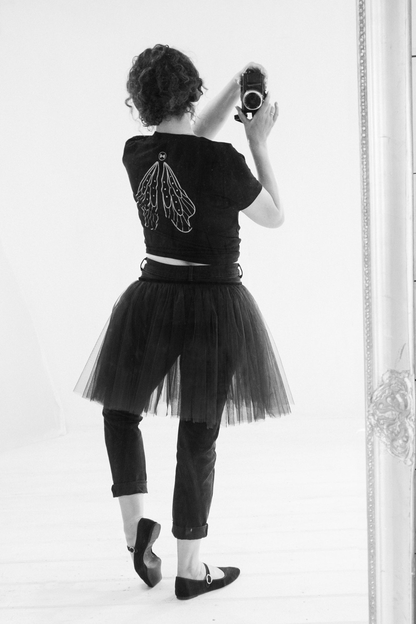 Person wearing a black tulle skirt and black crop top with a decorative fairy wing back design, standing in front of a mirror.