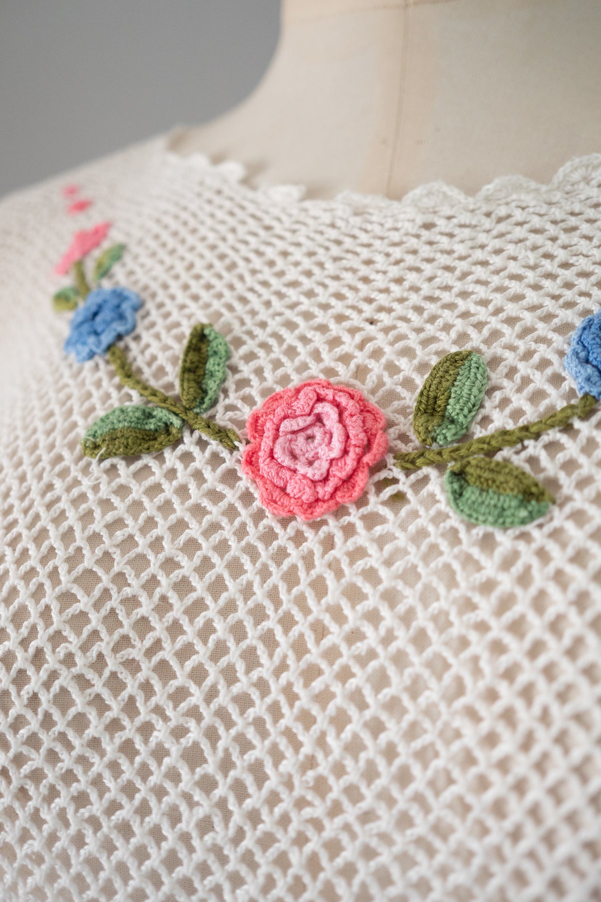 Close-up of a cream-colored crochet knit garment featuring a floral embroidered design with a prominent pink rose, green leaves, and blue flowers on the chest.