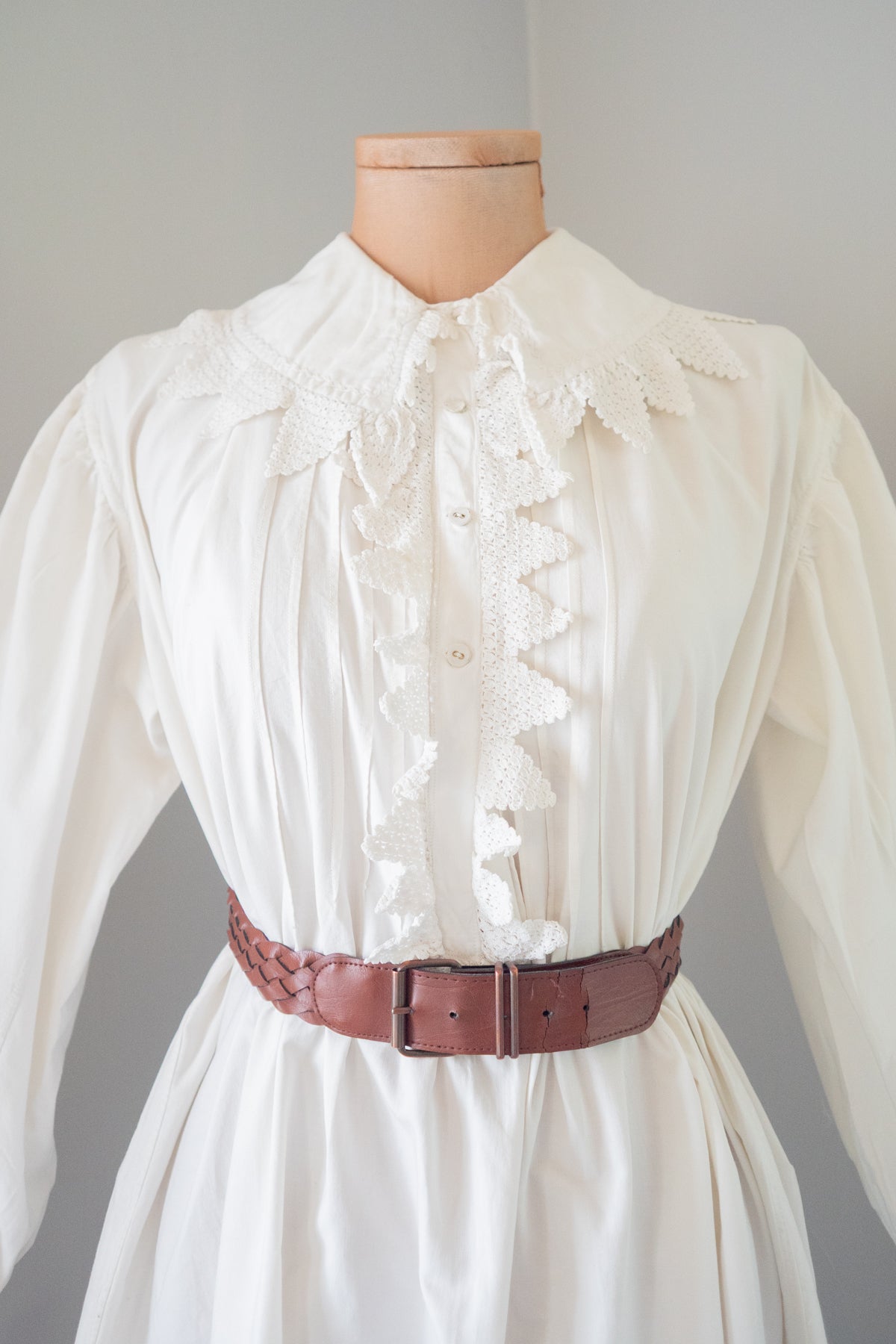 An antique white long-sleeve blouse or dress with a high lace collar and decorative ruffle placket, displayed on a mannequin torso and styled with a brown braided belt against a plain light gray background.