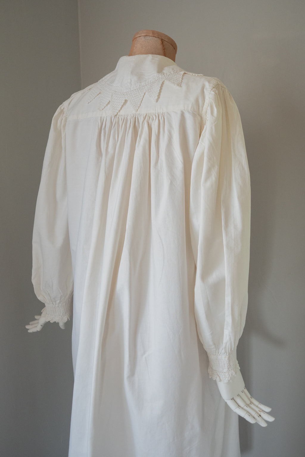 An off-white long-sleeve nightgown or smock with gathered shoulders and ruffled cuffs, displayed on a dress form mannequin against a plain gray wall.