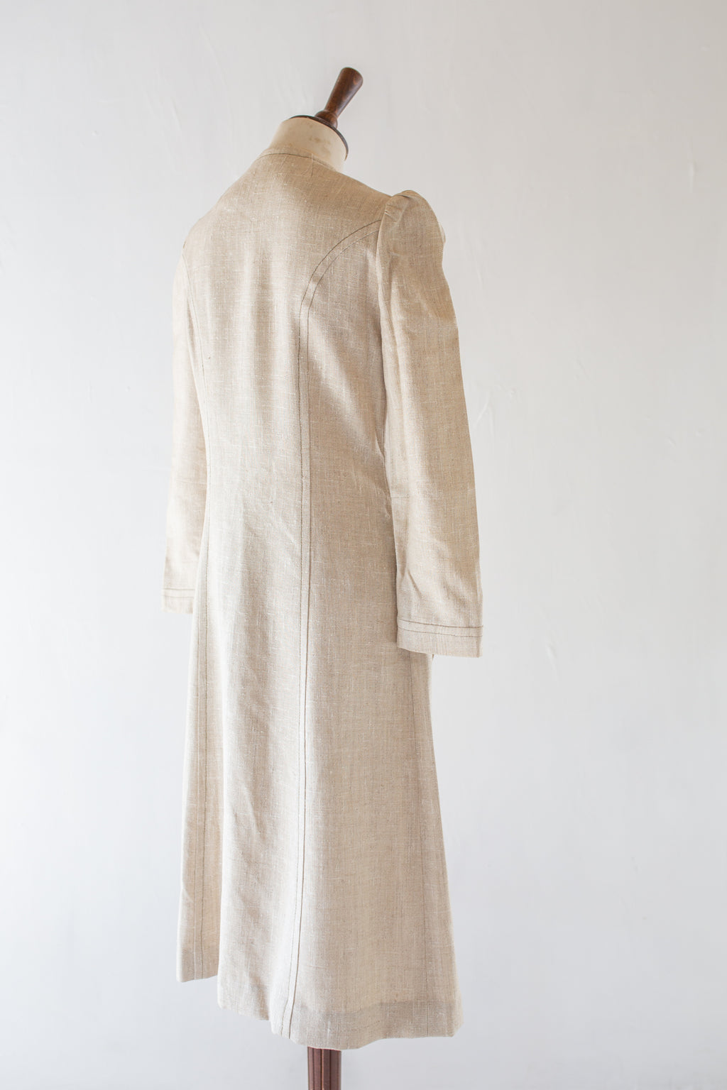 A light beige linen long coat displayed on a wooden dress form mannequin against a plain white background.
