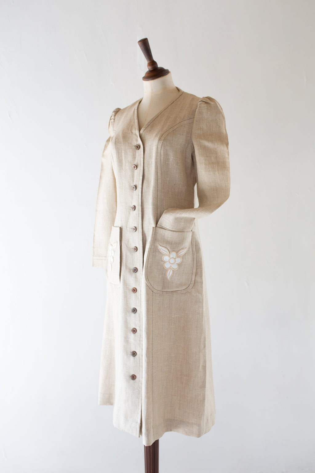 A vintage 1970s light beige linen coat dress with puff sleeves, a button-down front, and a decorative embroidered pocket, displayed on a wooden dress form against a white background.