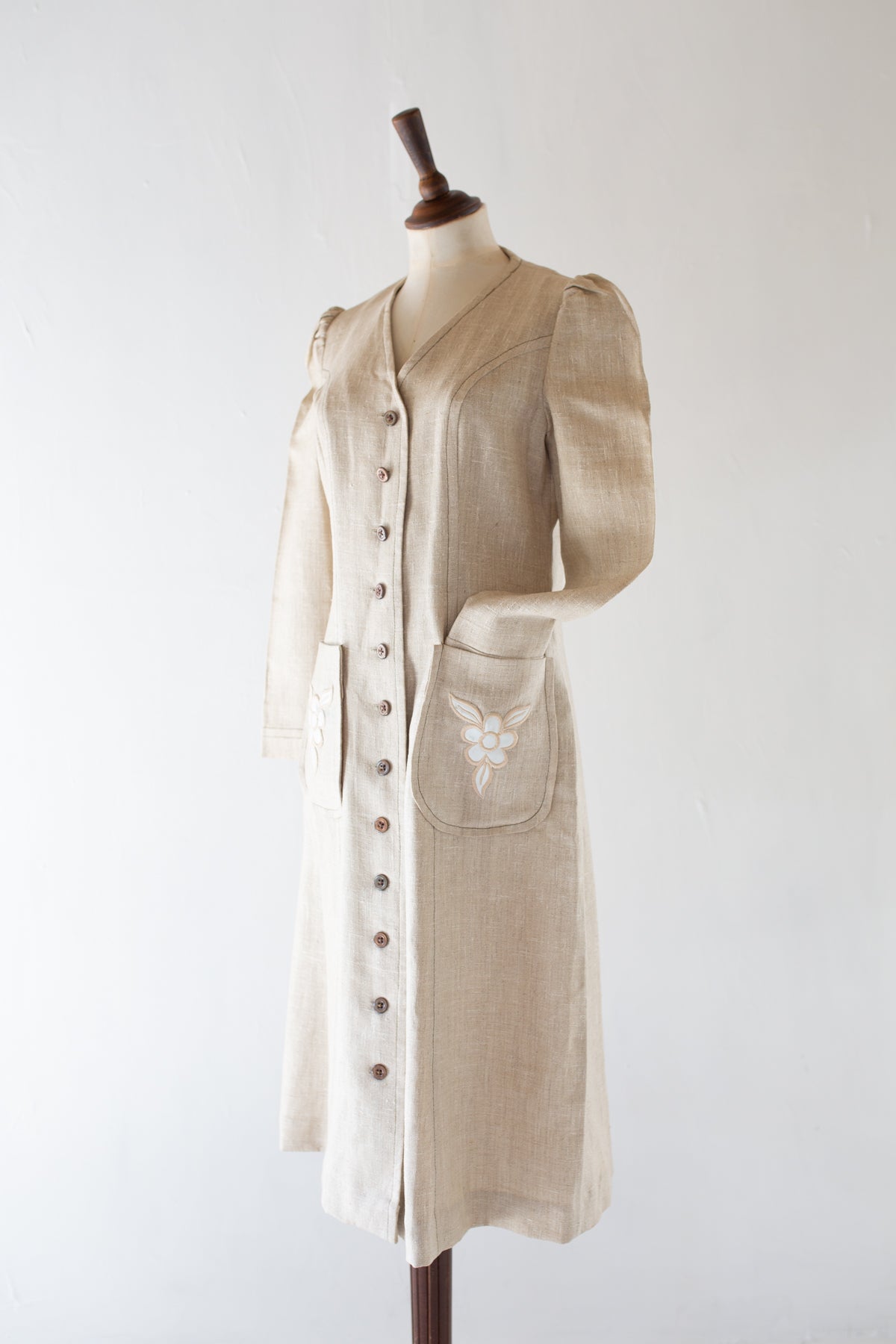 A vintage 1970s light beige linen coat dress with puff sleeves, a button-down front, and a decorative embroidered pocket, displayed on a wooden dress form against a white background.