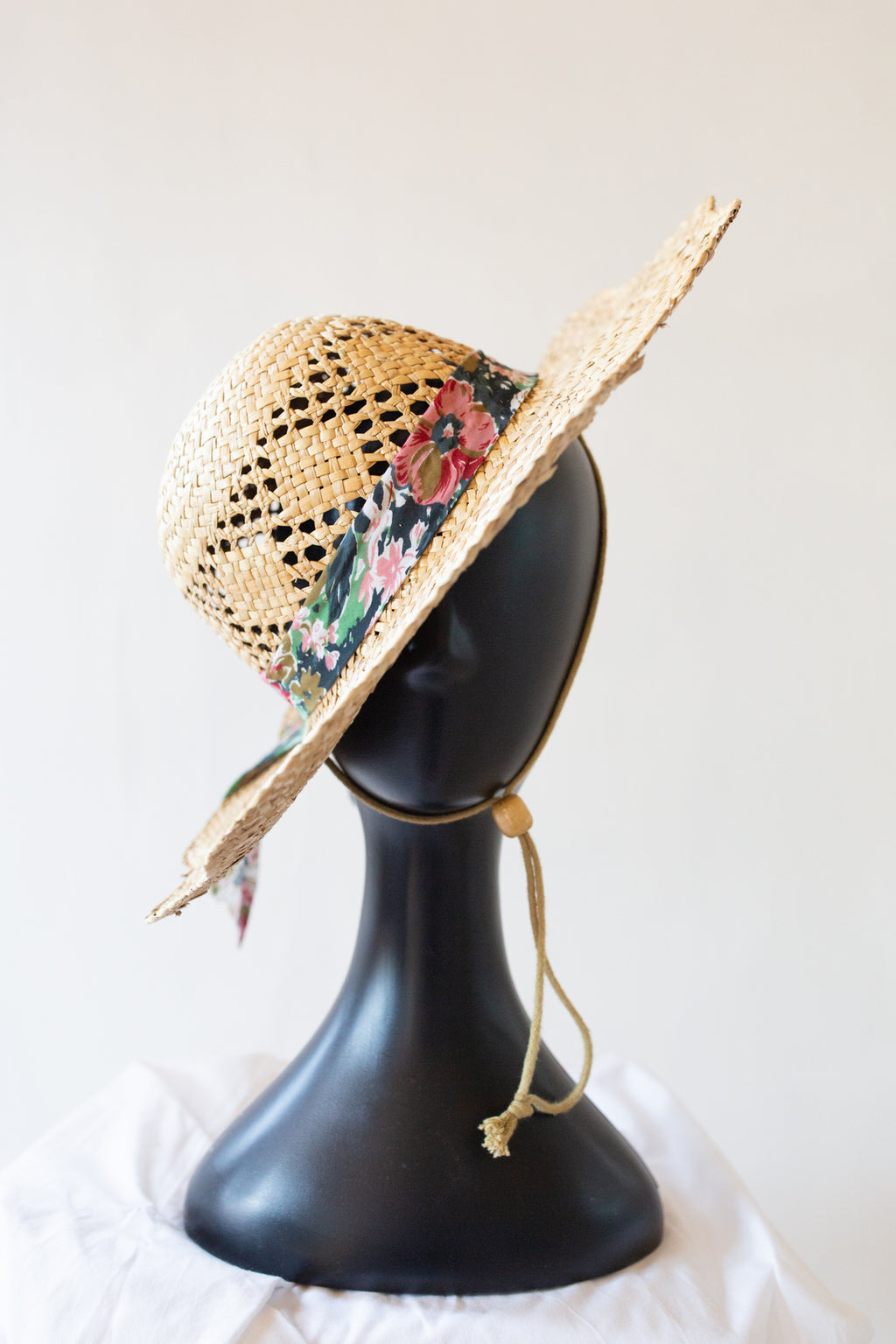 A straw sun hat with a wide brim, featuring a floral pattern band and a beige chin strap, displayed on a black mannequin head against a light background.