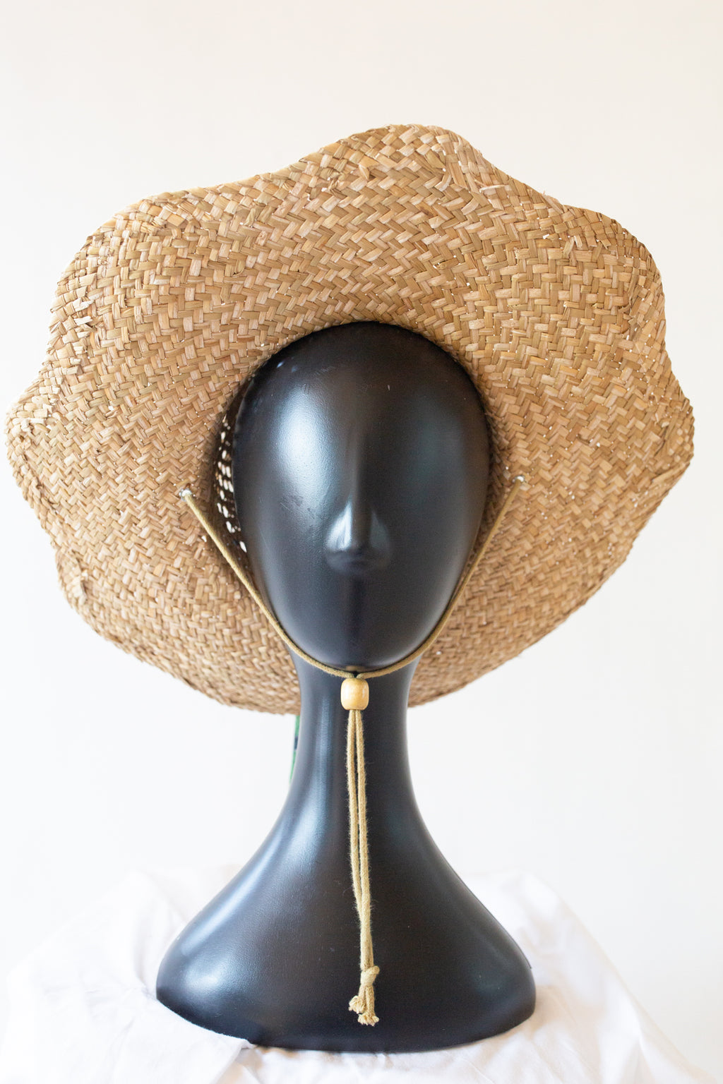 A natural woven straw sun hat with a wide brim and a tan chin cord, displayed on a black mannequin head against a plain white background.