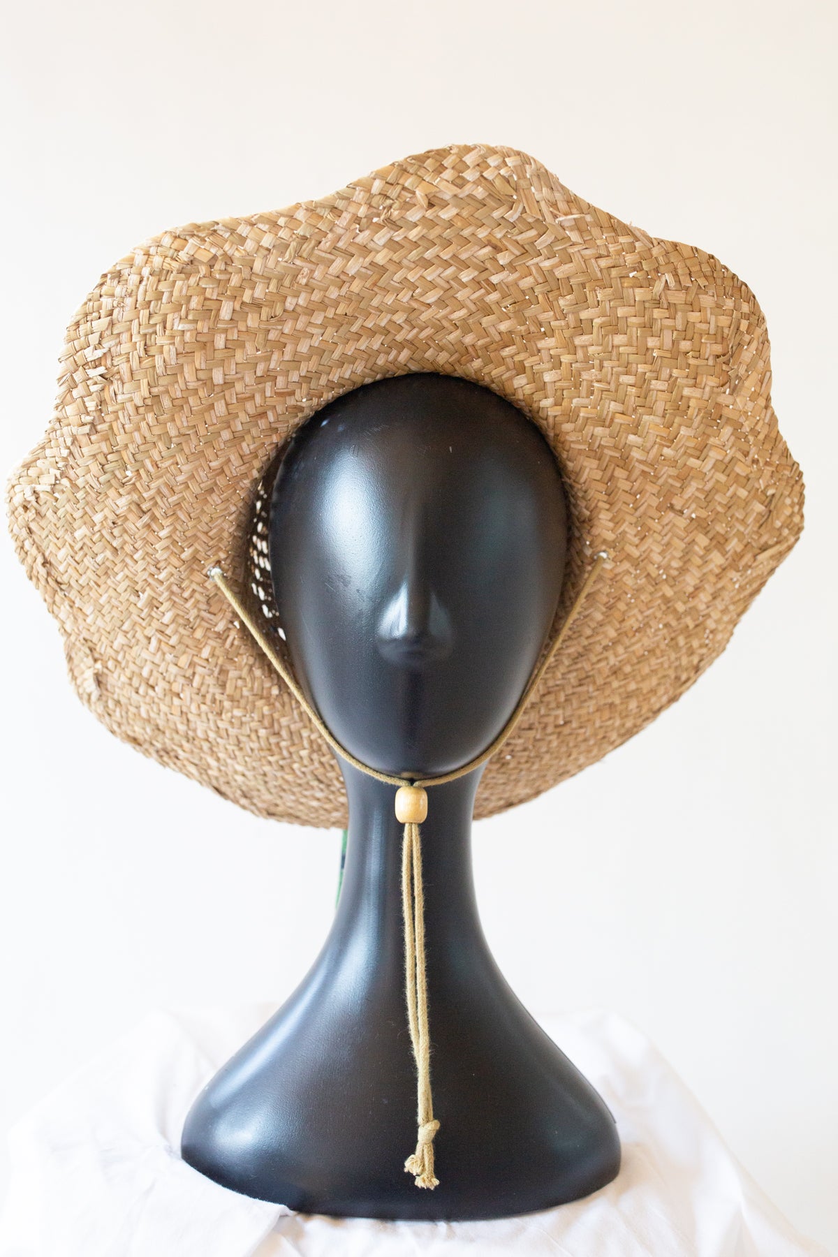 A natural woven straw sun hat with a wide brim and a tan chin cord, displayed on a black mannequin head against a plain white background.