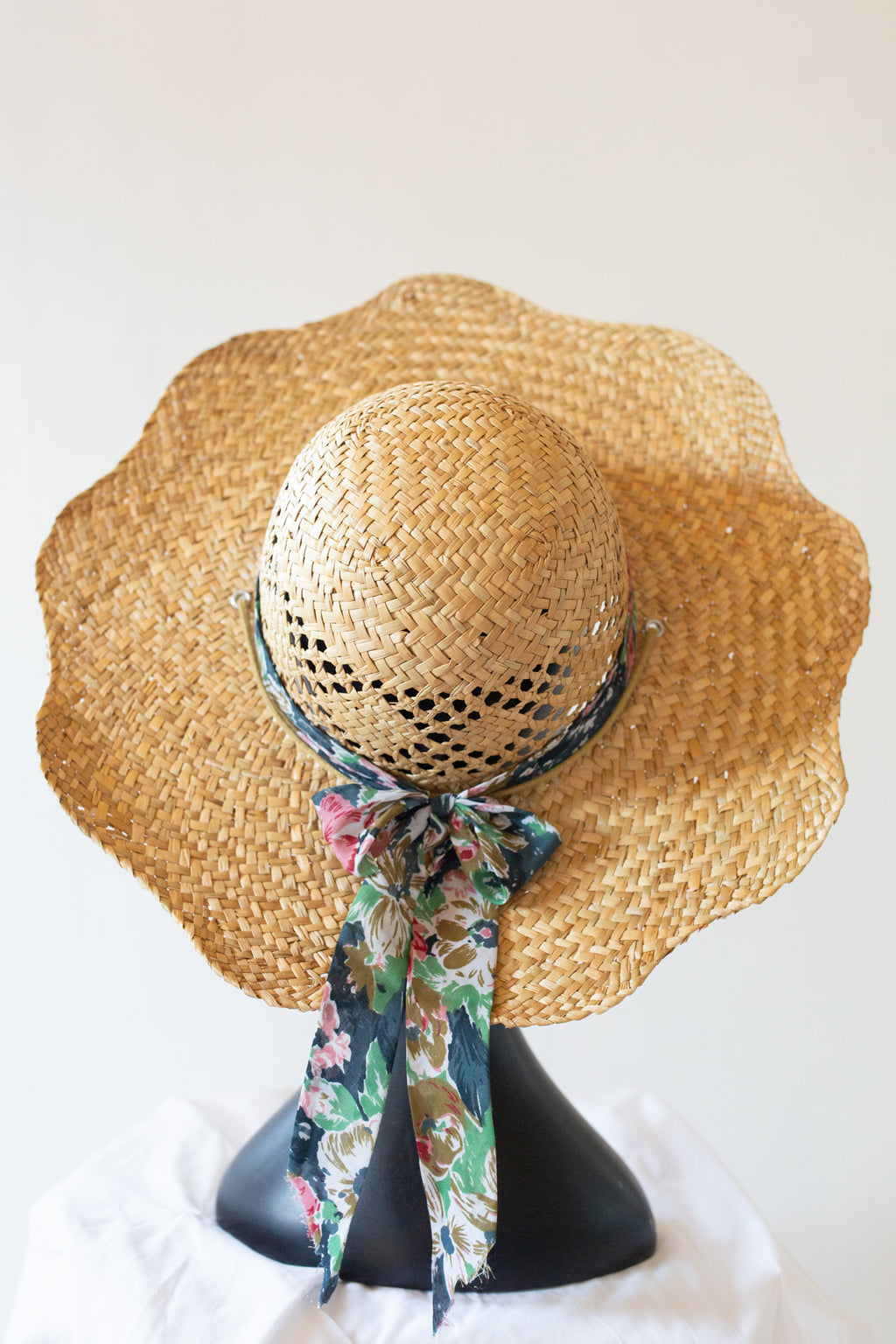 A vintage natural straw sun hat with a wide, scalloped brim and a dark patterned ribbon tied under the crown, displayed on a black mannequin head against a plain light background.