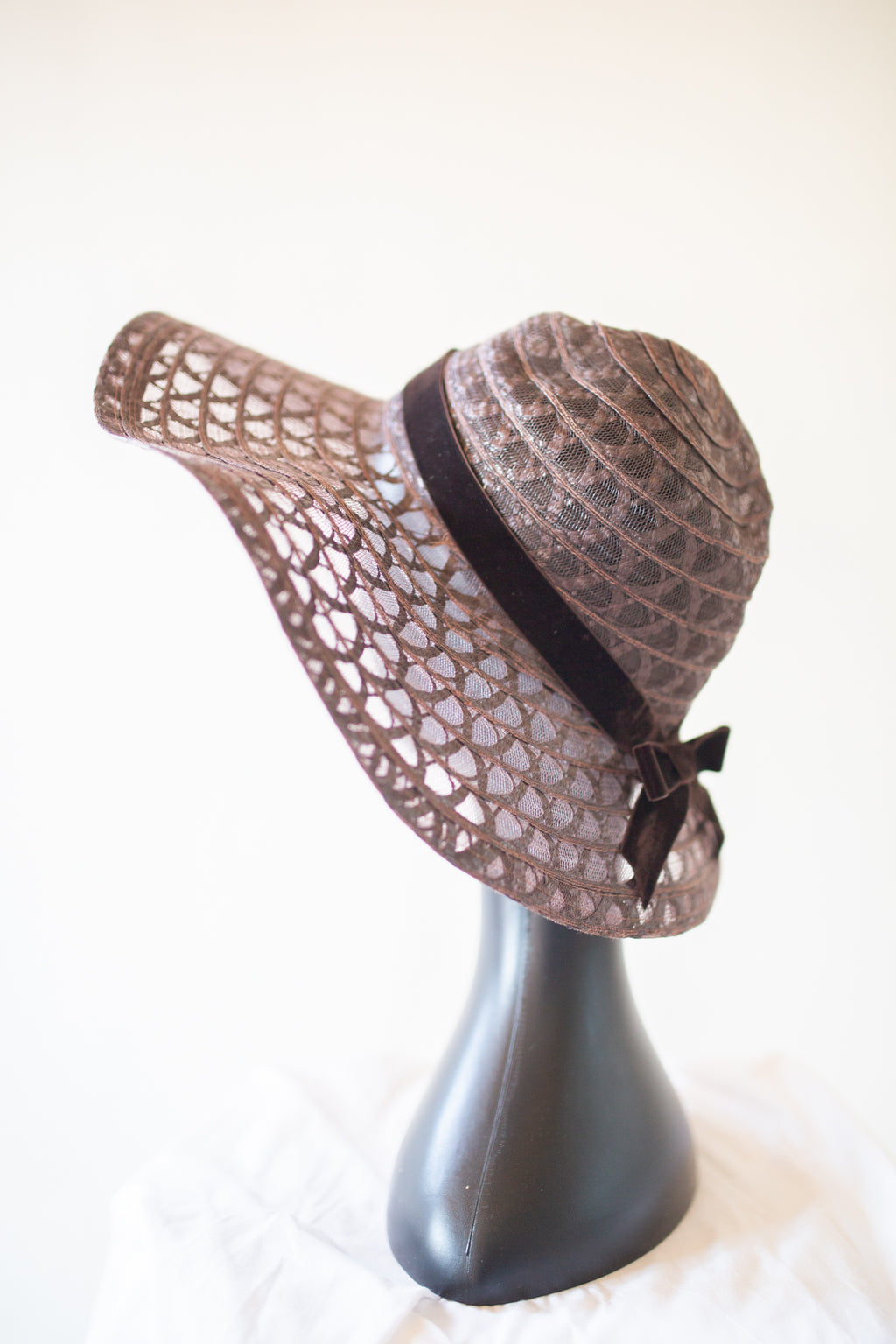 A brown woven sun hat with a wide brim, featuring a textured pattern and a dark brown ribbon band with a bow, displayed on a black mannequin head against a light background.