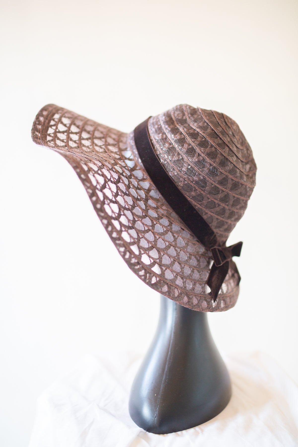 A brown woven sun hat with a wide brim, featuring a textured pattern and a dark brown ribbon band with a bow, displayed on a black mannequin head against a light background.