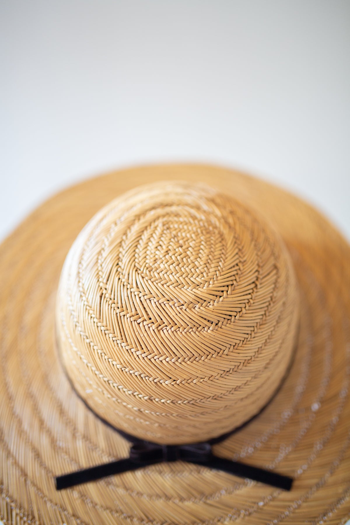 A close-up of a woven straw sun hat with a black ribbon bow, shown against a light gray background.