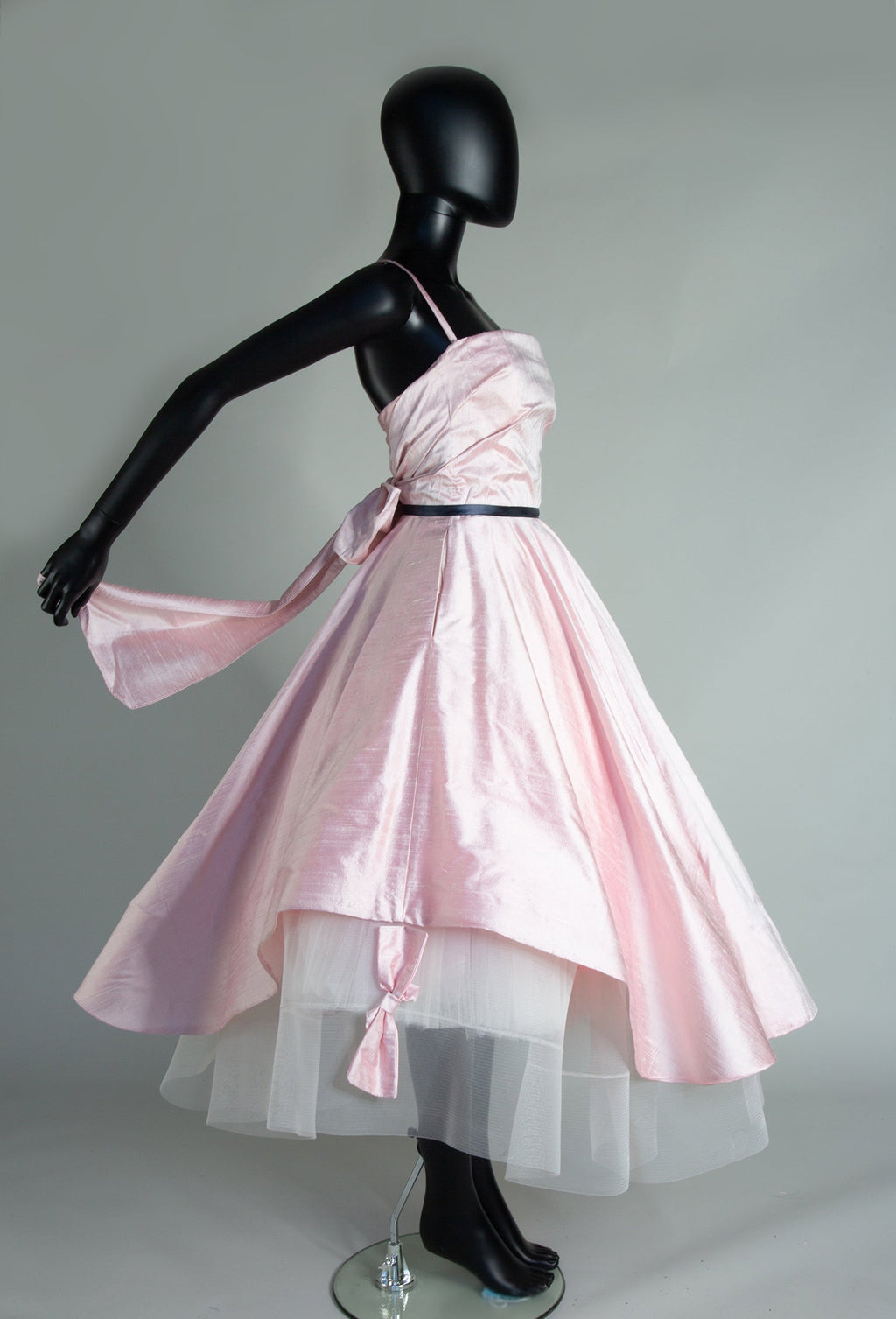 A mannequin displaying a peony blush pink silk gown with a fitted bodiceand a full circle skirt with silk bows.