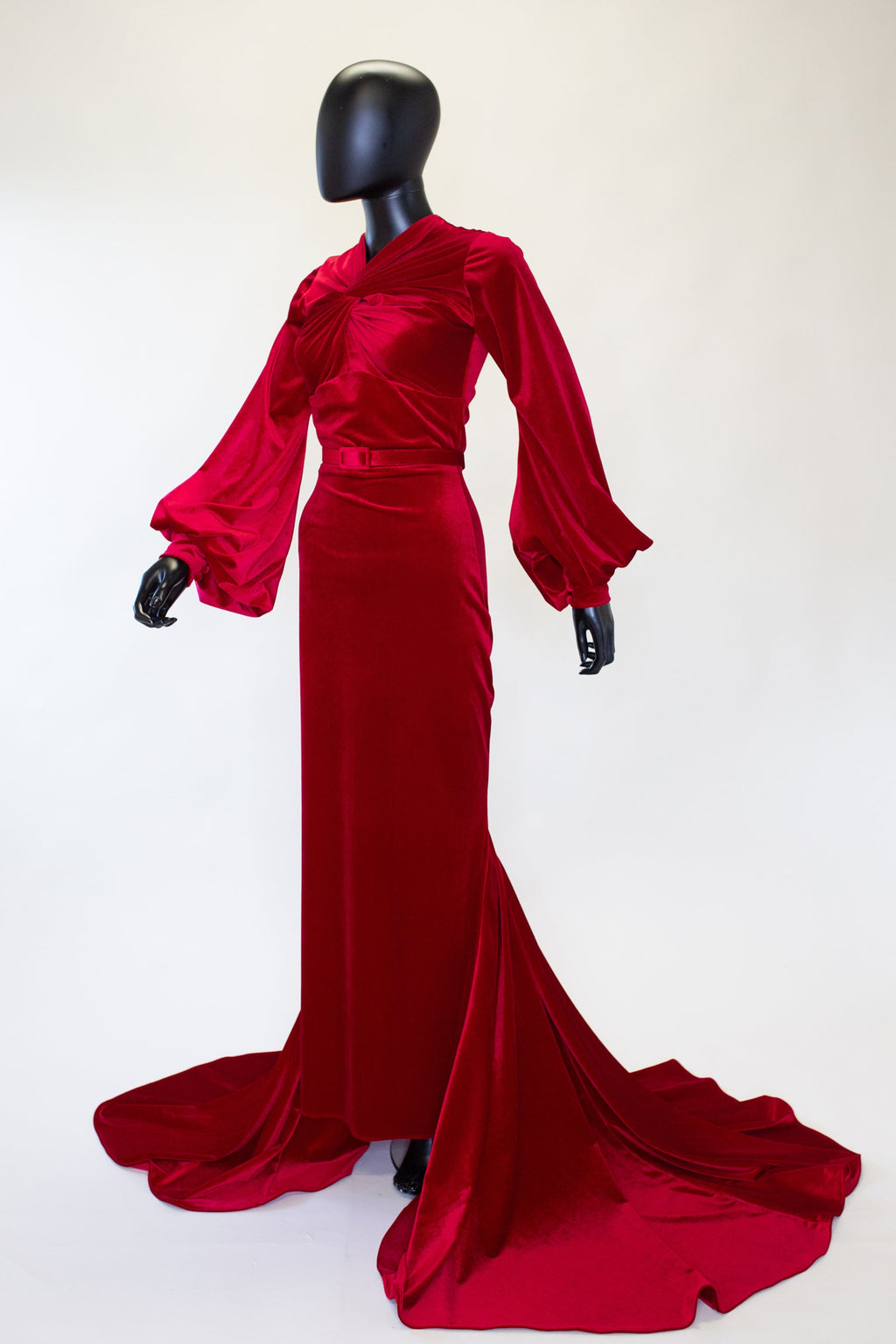 A red velvet old Hollywood gown with a twist bodice, waistline shaping, mermaid skirt, long fitted sleeves with buttons, and a puddle train at the back. It is displayed on a mannequin with no visible brand markings.